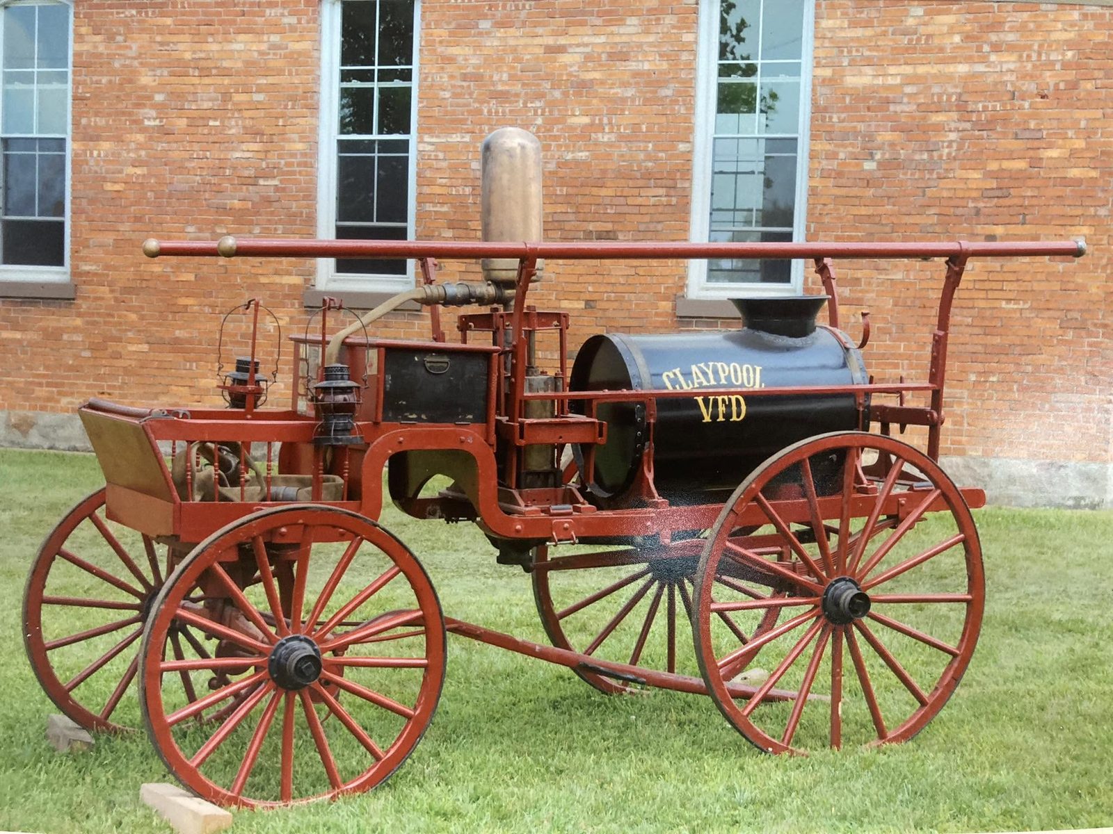 The department's restored horse-drawn Claypool VFD chemical engine, with red spoked wheels and a black tank lettered in gold, parked on the grass beside a brick building.